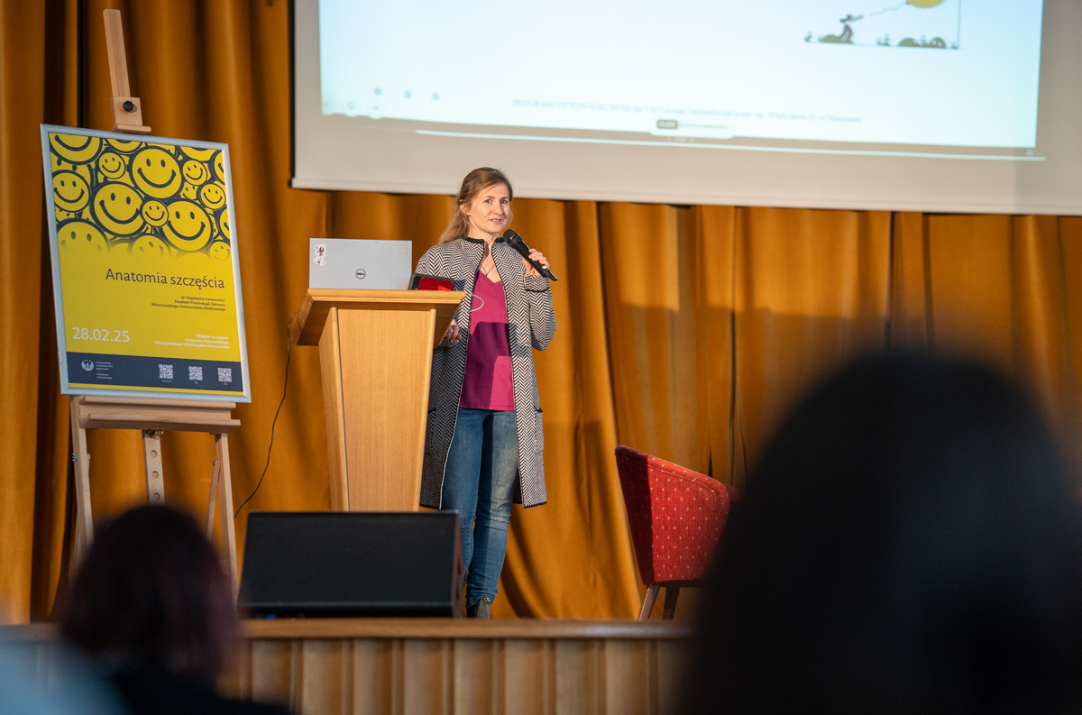 Osoba z mikrofonem stoi na scenie przed żółtą zasłoną, obok drewnianej mównicy z laptopem. Po lewej plakat z uśmiechniętymi buźkami i napisem „Anatomia szczęścia”. W tle ekran z prezentacją. Na pierwszym planie sylwetki dwóch osób z publiczności.