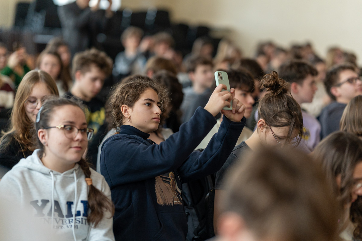 Grupa osób siedzi w sali. Na pierwszym planie osoba w ciemnej bluzie robi zdjęcie telefonem. W tle widać inne osoby i puste krzesła.