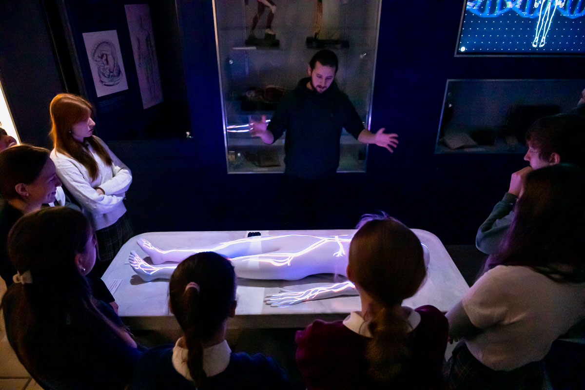 Grupa uczniów stoi wokół stołu z projekcją anatomicznego obrazu człowieka. Nauczyciel z rozłożonymi rękami tłumaczy coś grupie. W tle gabloty z eksponatami i ekran z grafiką DNA. Pomieszczenie jest ciemne, oświetlone głównie przez projekcję.