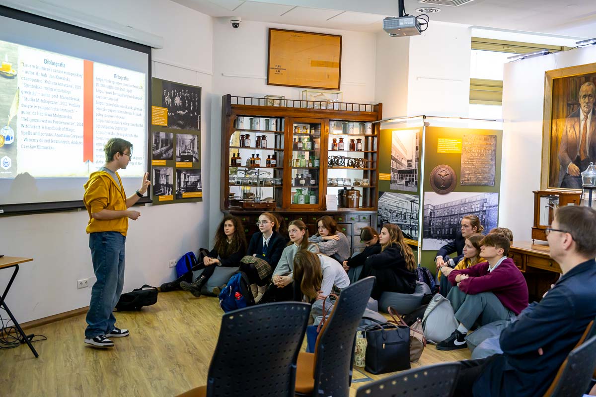 Mężczyzna w żółtej bluzie prowadzi prezentację w jasnym muzeum. Przed nim siedzi grupa osób na podłodze. Na ekranie widać slajd z tekstem i grafikami. W tle szklana witryna z eksponatami, plansze informacyjne, białe ściany i drewniana podłoga.