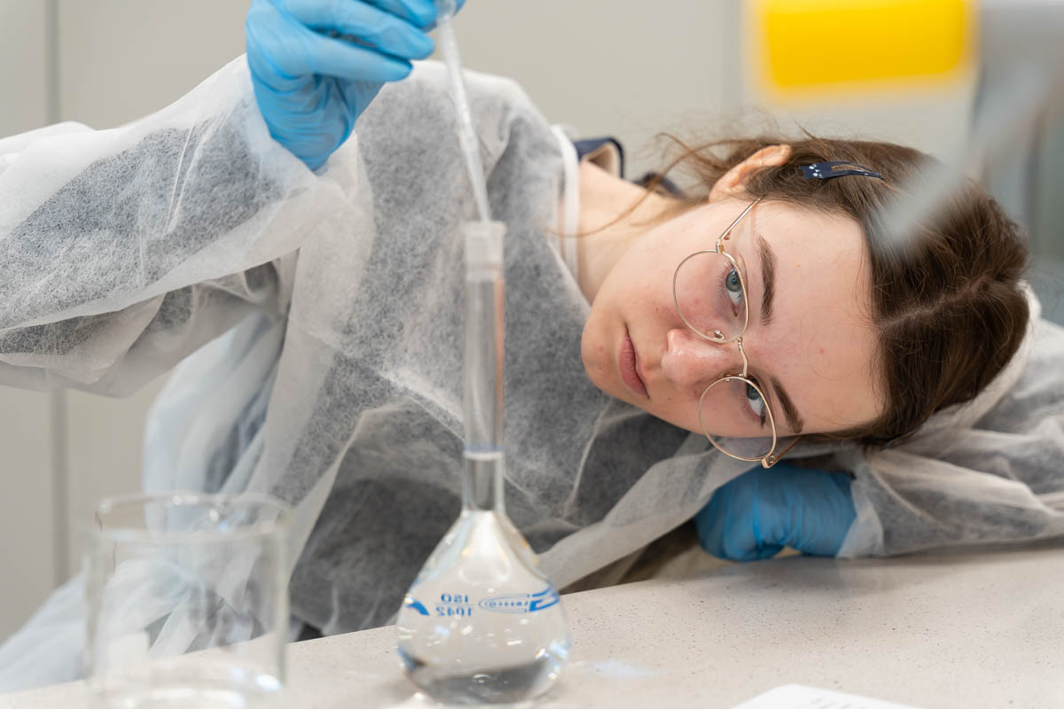 Osoba w białym fartuchu i niebieskich rękawiczkach przeprowadza eksperyment chemiczny. Pipetą przenosi ciecz do kolby stożkowej z przezroczystym płynem. Na stole stoi też szklanka laboratoryjna.