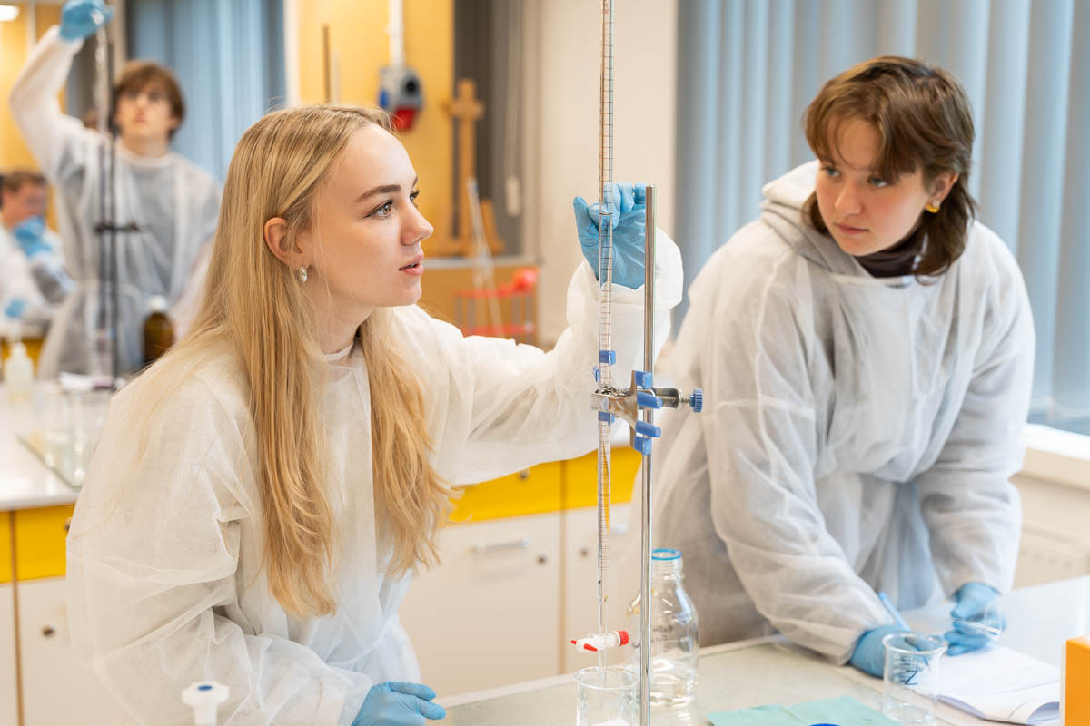 Dwie młode osoby w białych fartuchach i niebieskich rękawiczkach pracują w jasnym laboratorium chemicznym. Jedna trzyma biuretę, druga obserwuje. W tle widać sprzęt, inne osoby, żółte szafki i duże okna z niebieskimi żaluzjami.