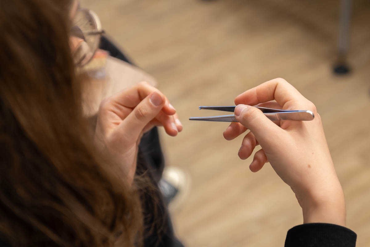 Osoba trzyma pęsetę w prawej ręce i przygląda się jej z bliska. Lewa ręka jest uniesiona, jakby miała coś chwycić. W tle rozmyta drewniana podłoga.