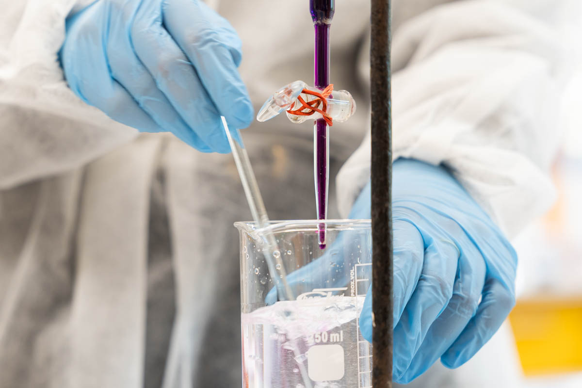 Osoba w białym fartuchu i niebieskich rękawiczkach trzyma pipetę nad cylindrem miarowym z przezroczystą cieczą. Cylinder ma podziałkę do 50 ml. Scena przedstawia eksperyment chemiczny.