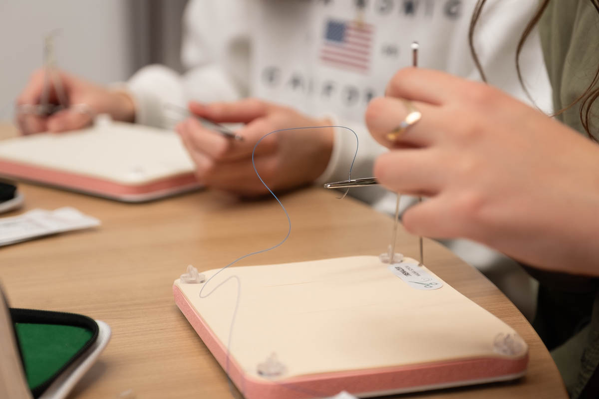 Dwie osoby ćwiczą szycie chirurgiczne na symulatorach. Na drewnianym stole leżą podkładki z materiałem imitującym skórę, nici i narzędzia. Osoba po prawej trzyma igłę z nicią, osoba po lewej używa narzędzi.