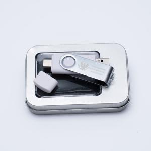 Biało-szary pendrive leżący na srebrnym pudełku. Pendrive ma zdjętą zatyczkę osłaniającą wtyk.