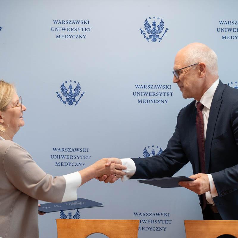 Kobieta, dyrektor NIO i mężczyzna, rektor WUM stoją naprzeciwko siebie i podają sobie ręce. Jedna ubrana w beżowy sweter, druga w ciemny garnitur z białą koszulą i bordowym krawatem. W tle niebieska ściana z logo i napisem „Warszawski Uniwersytet Medyczny”. W rękach mają dokumenty. Kobieta jest ubrana w jasnym sweter, mężczyzna w ciemny garnitur.