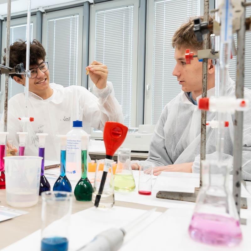 Dwaj młodzi mężczyźni w białych fartuchach laboratoryjnych siedzą przy stole w laboratorium farmaceutycznym. Przed nimi są kolorowe barwniki, mezurki i przyrządy farmaceutyczne.